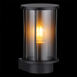 Moderne zwarte wandlamp voor buiten met cilindervormig design en helder glas, frontaal schuin aanzicht met zichtbare filamentlamp in warme gloed.