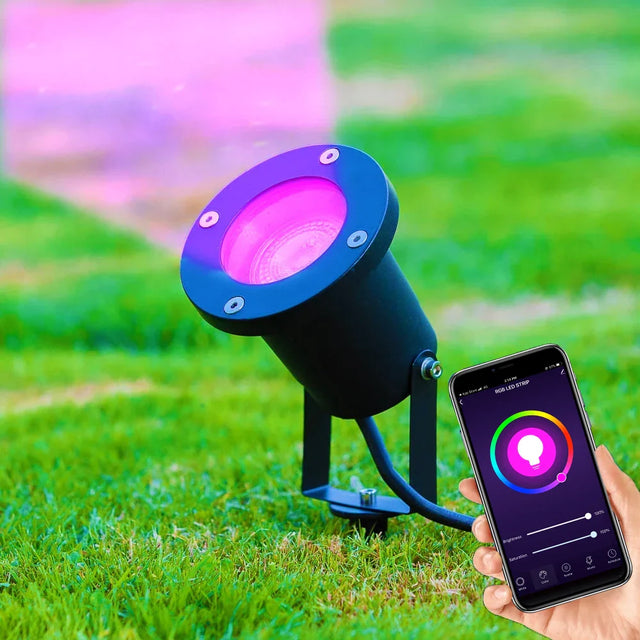 Zwarte slimme RGB LED-buitenspot op gras, met roze lichtstraal, bediend via smartphone-app – zijaanzicht.