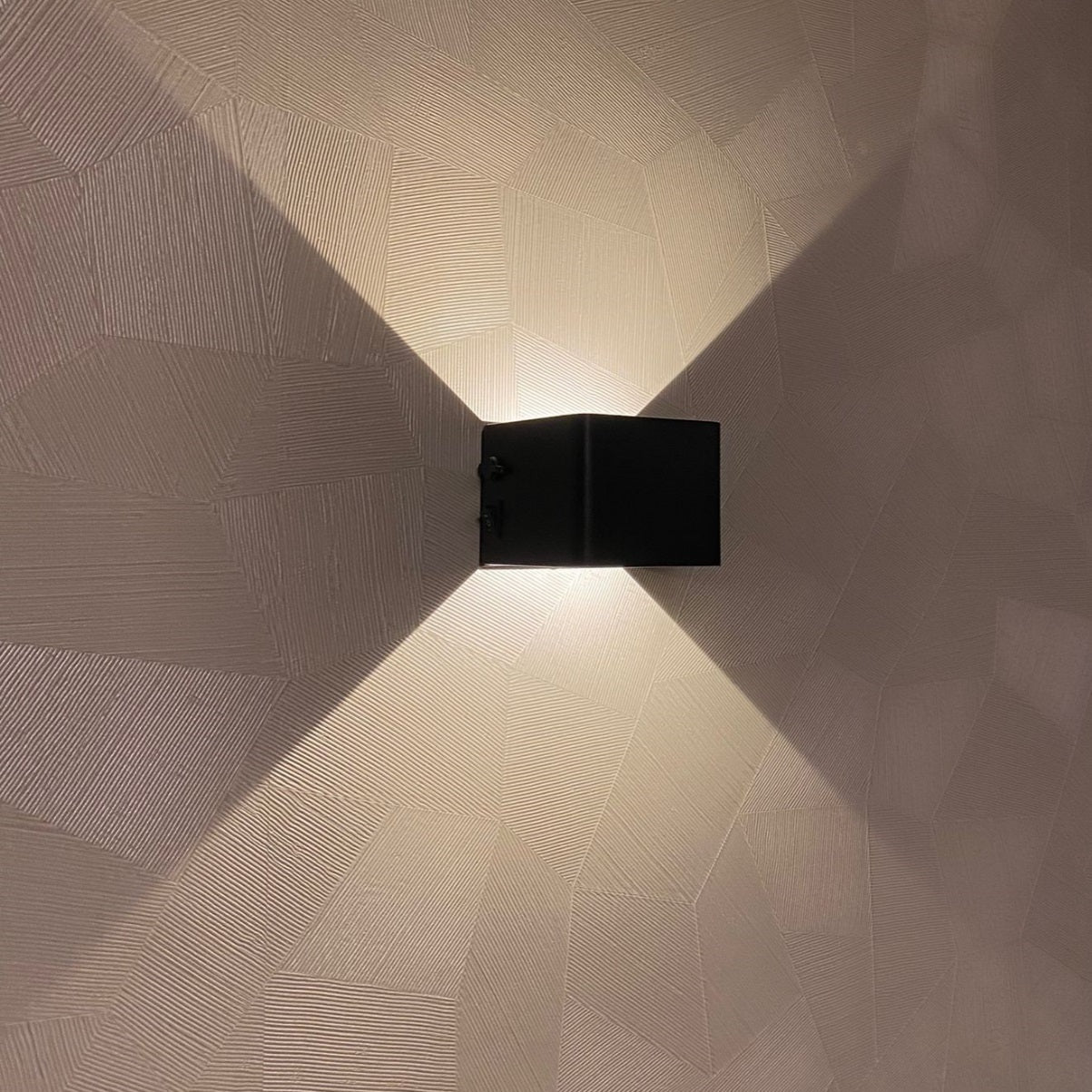 Moderne zwarte wandlamp met op- en neerwaartse lichtbundels, gemonteerd op een decoratief gestucte muur – vooraanzicht.