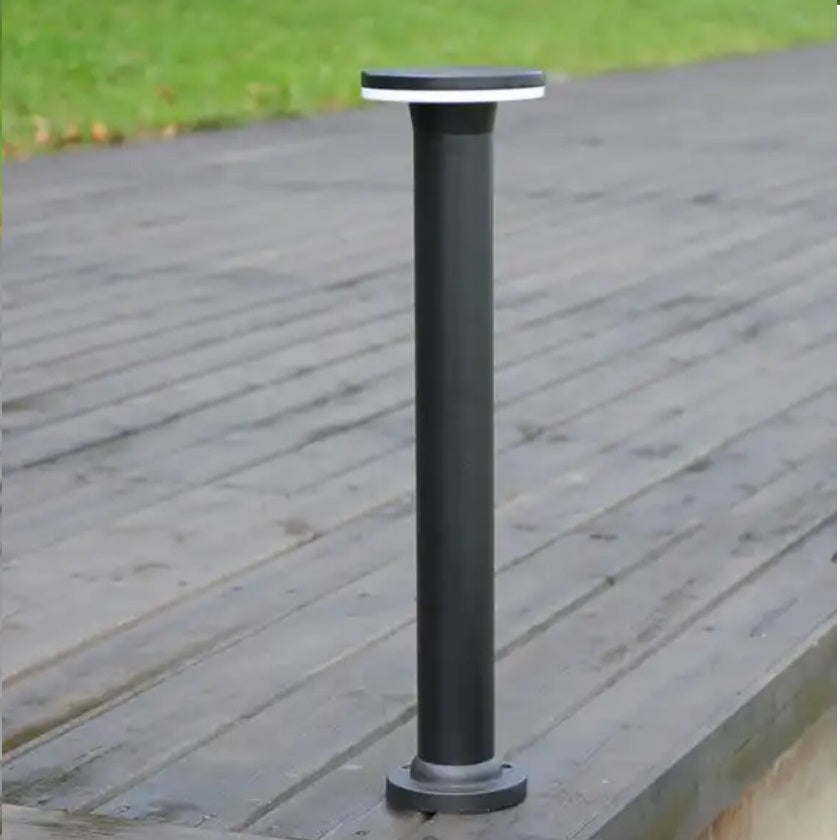 Zwarte moderne buitenlamp op zonne-energie in staande sokkelvorm, gefotografeerd in perspectivisch zijaanzicht op een houten terras met gras op de achtergrond.