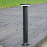 Zwarte moderne buitenlamp op zonne-energie in staande sokkelvorm, gefotografeerd in perspectivisch zijaanzicht op een houten terras met gras op de achtergrond.