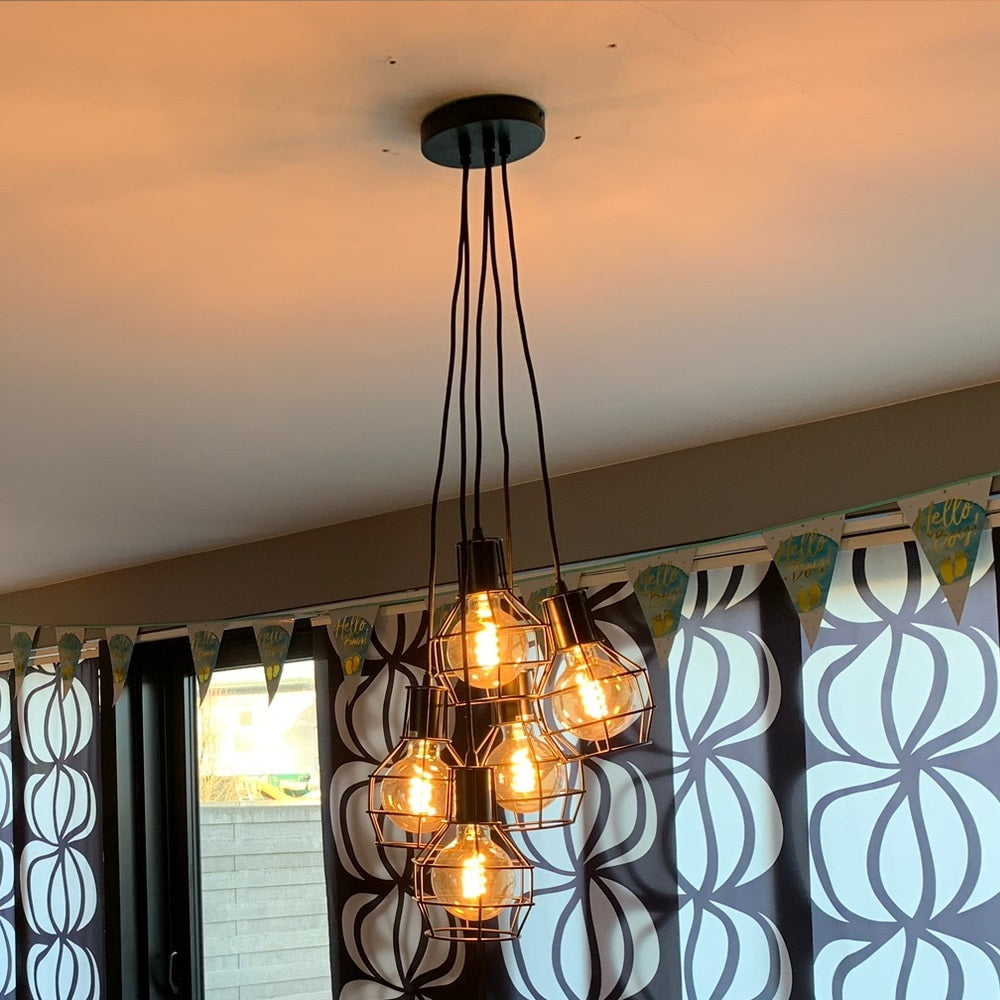 Industriële zwarte hanglamp met zes vintage filament LED-lampen in metalen draadframes, schuin van onderen gefotografeerd tegen een plafond met lichtweerkaatsing in een modern interieur.