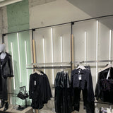 Winkelinterieur met kledingdisplay tegen een wand met verticale LED-verlichting; vooraanzicht van zwarte dameskledingstukken, waaronder jurken, broeken, tops en een leren jas op een mannequin.