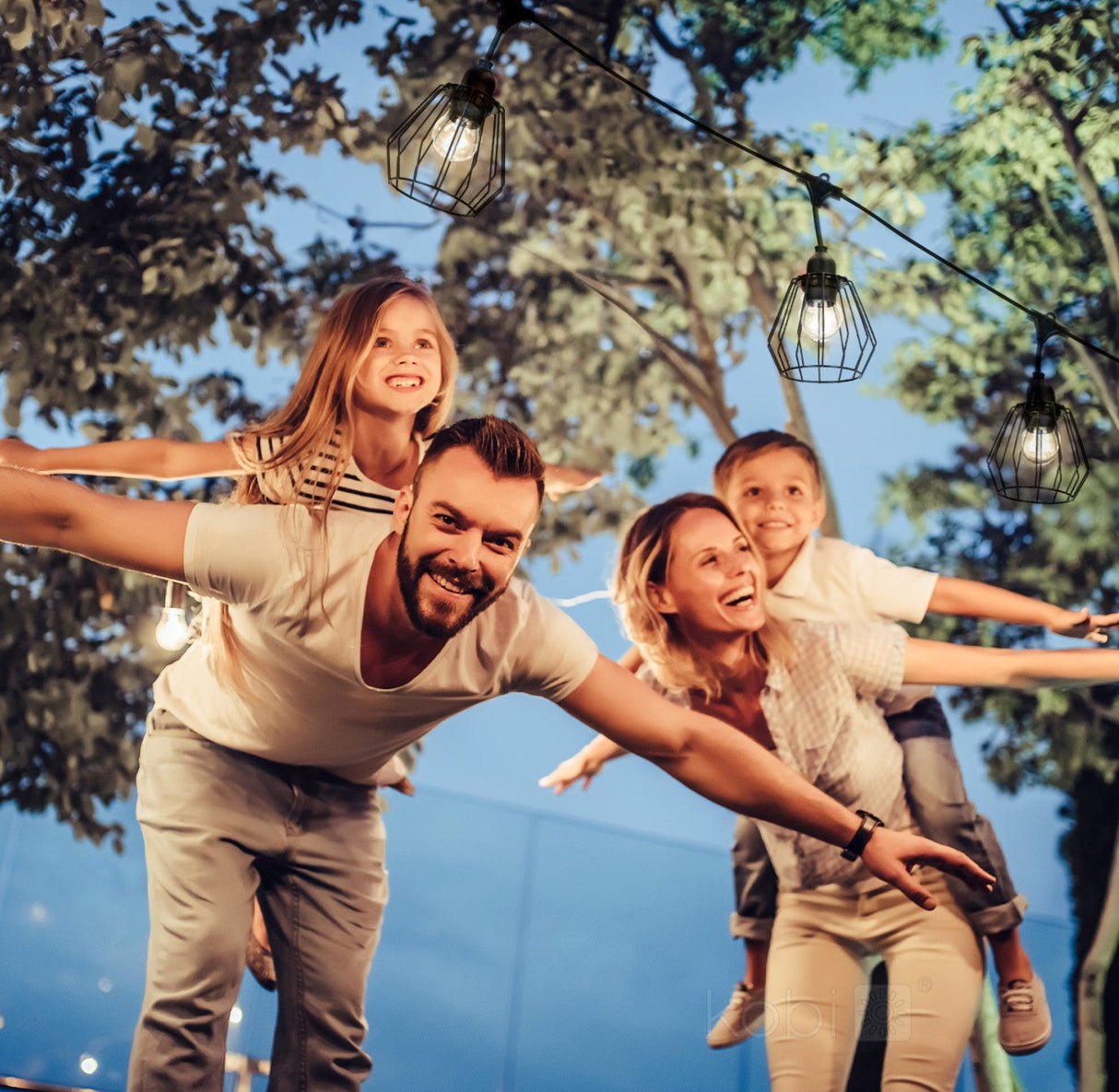Blije familie geniet van een zomerse avond in de tuin, verlicht door decoratieve buitenverlichting met retro kooi-achtige lampenkappen – vooraanzicht, met focus op de sfeerverlichting en lachende gezichten.