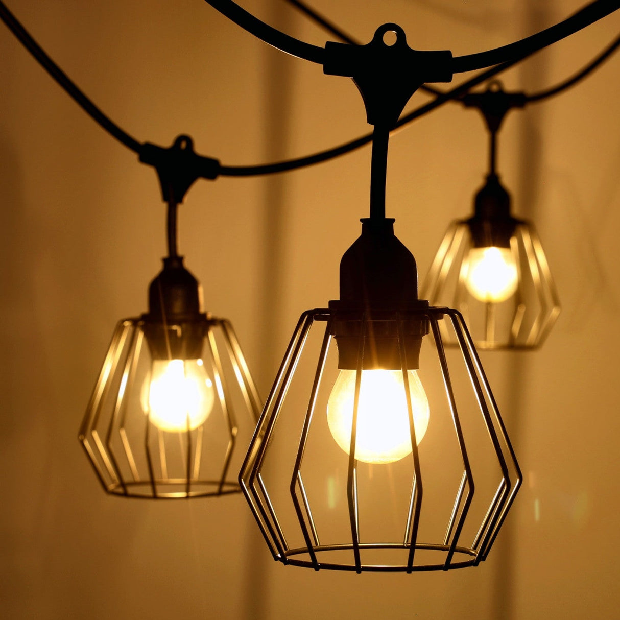 Close-up van industriële stringlight buitenlampen met metalen kooikap en warme LED-verlichting, hangend in een sfeervolle avondsetting.