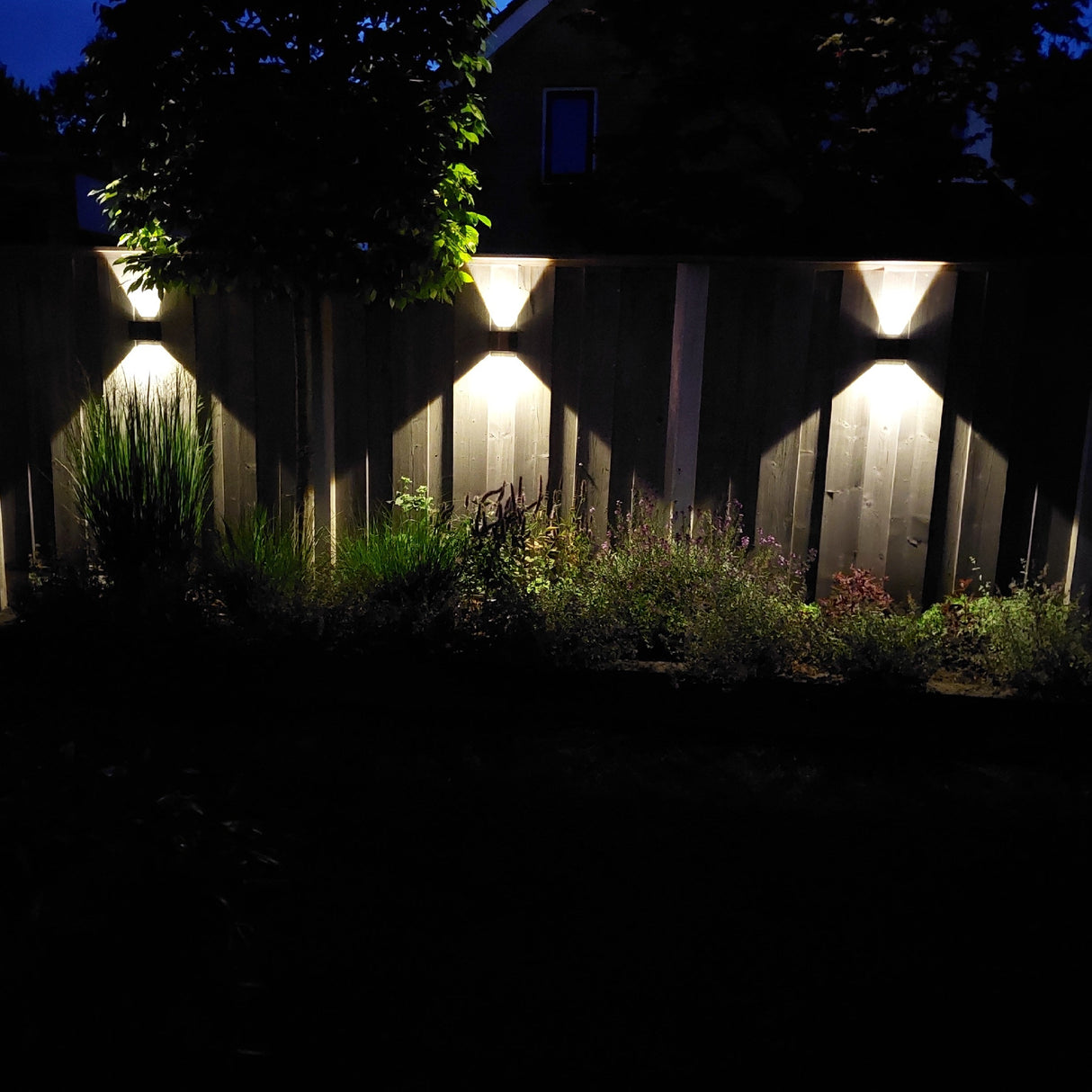 Moderne buitenwandlampen met up- en downlight verlichting gemonteerd op een houten schutting, verlichtend effect in de vorm van een X zichtbaar in nachtelijke tuinsetting, vooraanzicht.