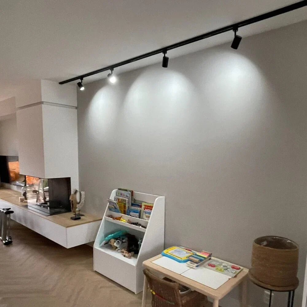 Kinderboekenkast in witte kleur, schuin vooraanzicht, geplaatst in een moderne woonkamer met houten vloer, naast een kindertafel en onder een zwarte plafondspotverlichting.