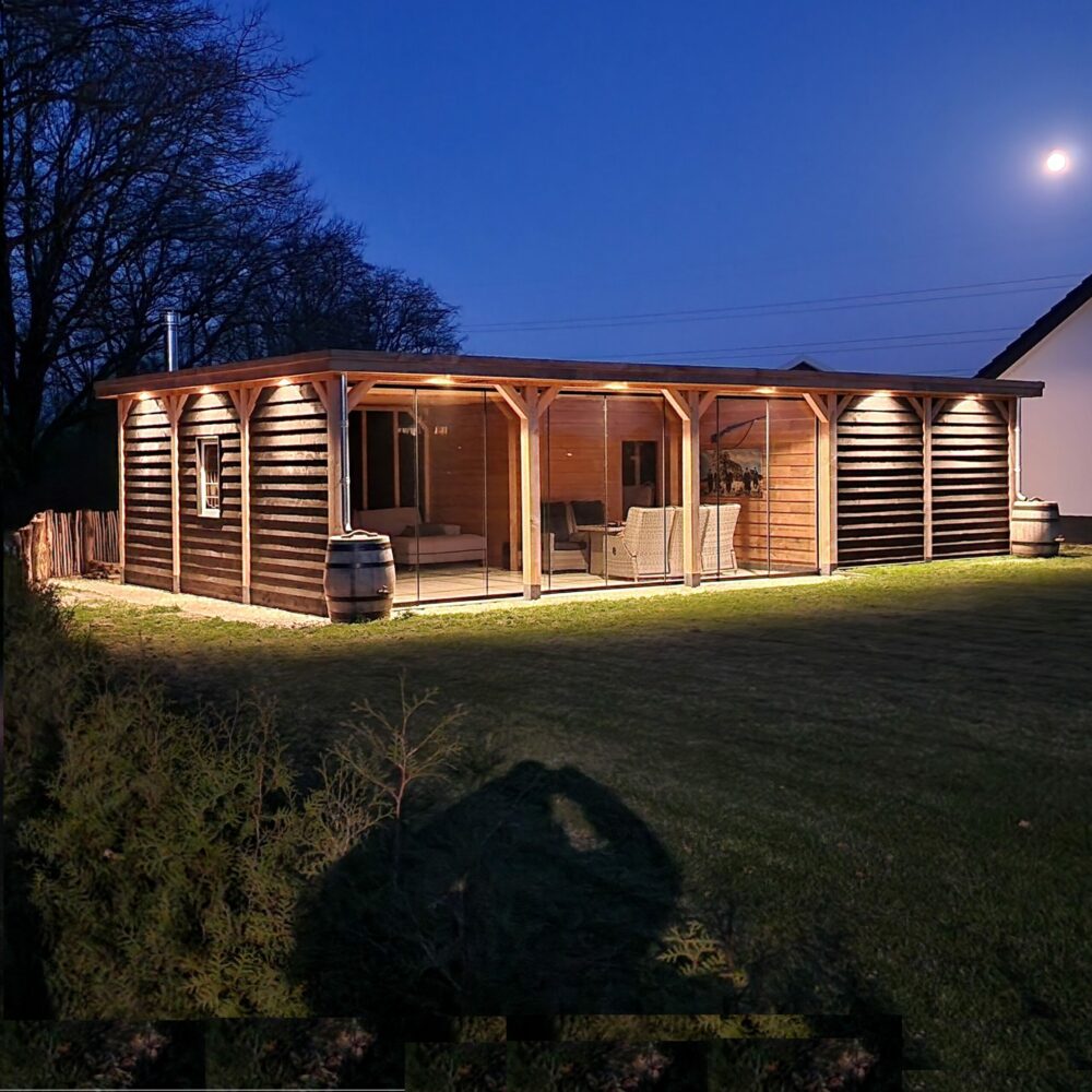 Houten overkapping met glazen schuifwanden en geïntegreerde verlichting, sfeervol verlicht in de avond. Schuin vooraanzicht met zicht op terras, tuinmeubilair en houten gevelbekleding.