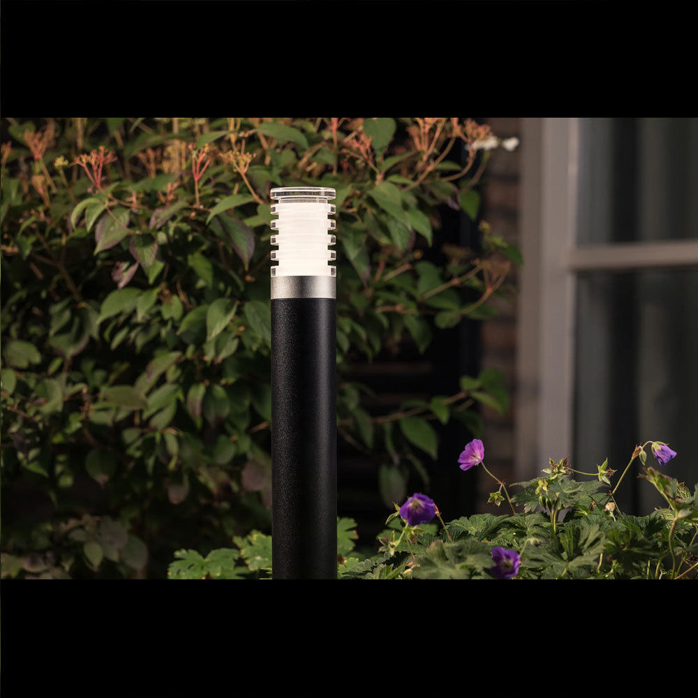 Moderne zwarte buitenlamp met RVS accenten en transparante lamellenkap, staand in een tuin met bloemen en groen blad, gefotografeerd in een frontaal aanzicht bij schemering.