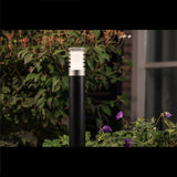 Moderne zwarte buitenlamp met RVS accenten en transparante lamellenkap, staand in een tuin met bloemen en groen blad, gefotografeerd in een frontaal aanzicht bij schemering.