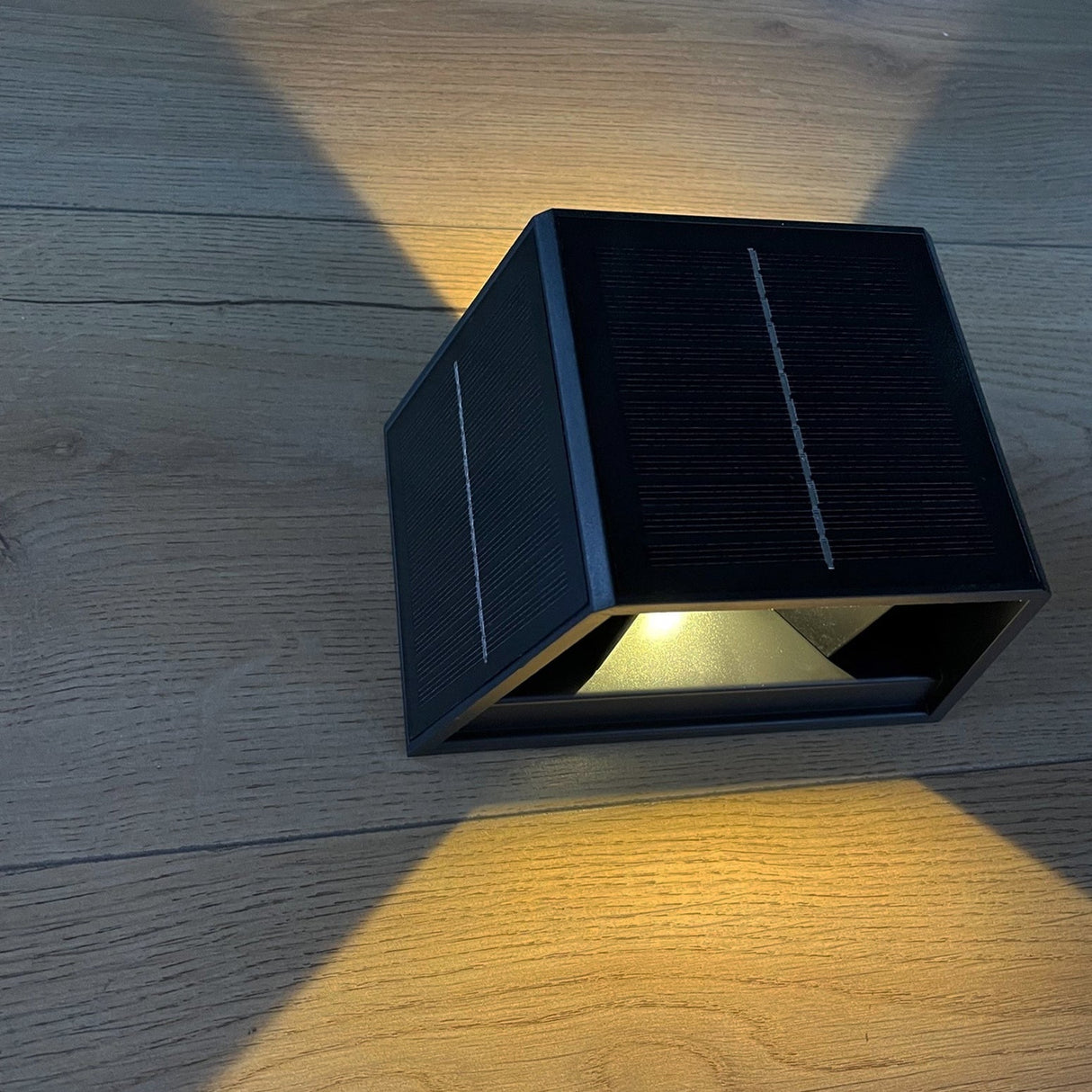 Zwarte solar wandlamp met moderne driehoekige vorm, gemonteerd op houten ondergrond, met zichtbare boven- en zijpanelen voorzien van zonnecollectoren en warm wit lichtstraal zichtbaar aan de onderzijde – schuin bovenaanzicht.