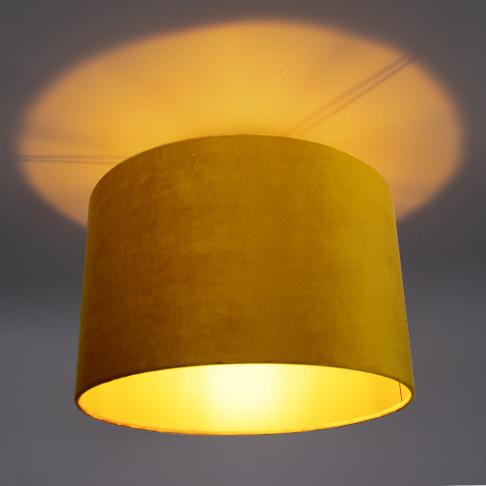 Gele velvet plafondlamp met cilinder vorm, gezien van schuin onderaanzicht, met warm licht dat een zachte gloed op het plafond werpt.