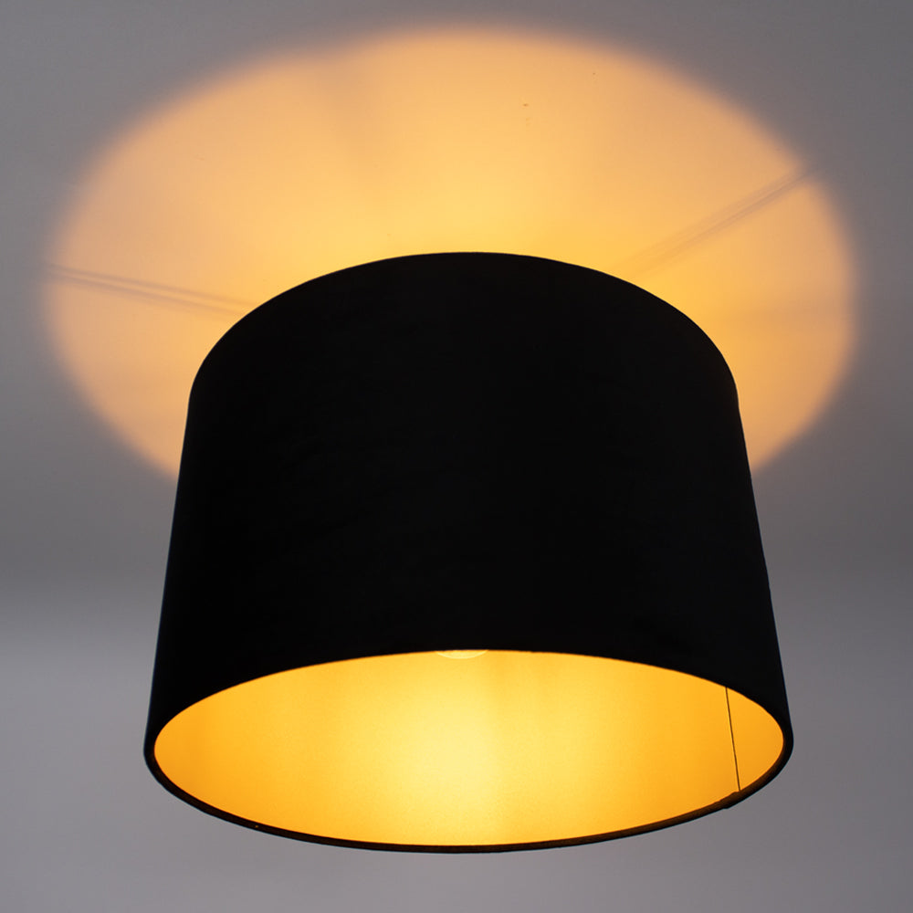 Zwart-gouden plafonnière met cilinderkap, gezien vanaf de onderzijde, met warme, sfeervolle lichtuitstraling tegen een witte plafondachtergrond.