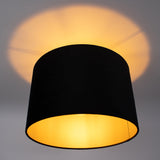 Zwart-gouden plafonnière met cilinderkap, gezien vanaf de onderzijde, met warme, sfeervolle lichtuitstraling tegen een witte plafondachtergrond.