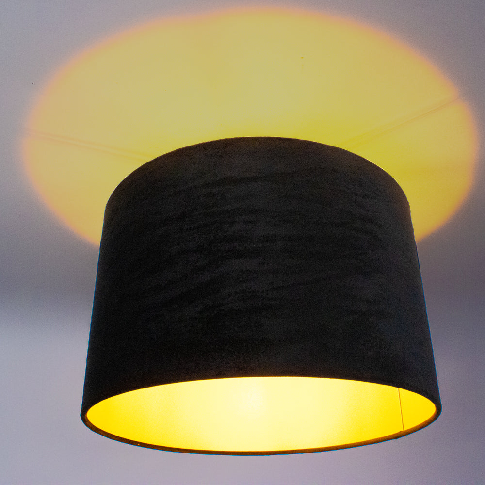 Zwarte plafondlamp met cilindervormige kap, binnenzijde goudkleurig, verspreidt warm licht tegen het plafond – onderaanzicht.