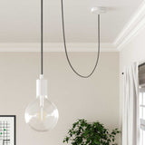 Minimalistische hanglamp met zwarte stoffen kabel en witte fitting, met zicht op de zijkant in een modern interieur met lichte muren, een kamerplant en een raam met gordijn.