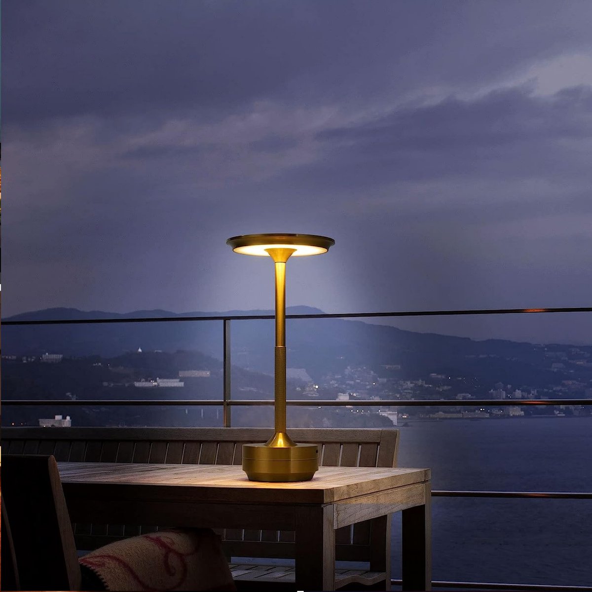 Gouden tafellamp met modern design op houten buitentafel, verlicht in schemering met uitzicht op berglandschap en zee – zijaanzicht.