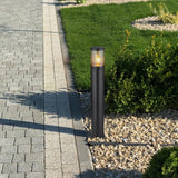 Staande buitenlamp met moderne zwarte afwerking, geplaatst langs een tuinpad met grind en gras; frontaal zijaanzicht tijdens daglicht met warme LED-verlichting zichtbaar.