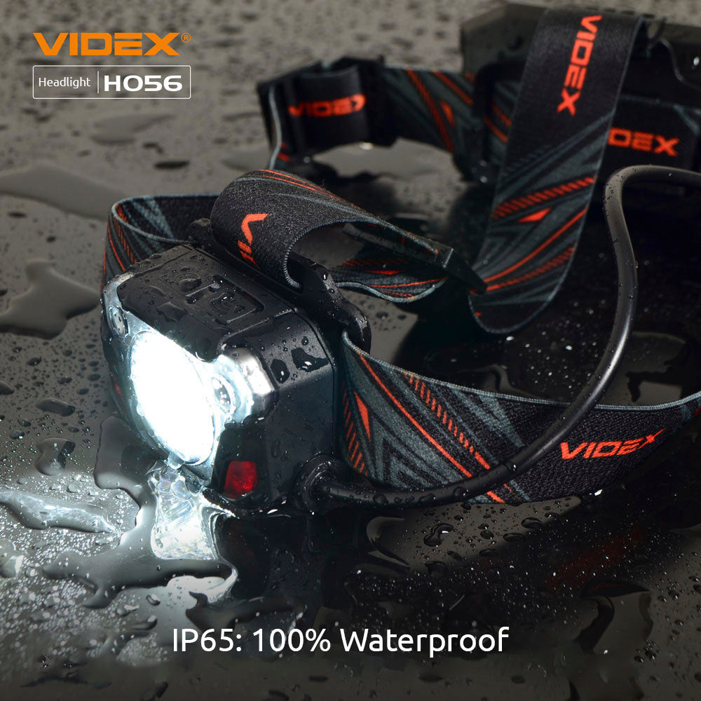 Close-up zijaanzicht van de Videx HO56 hoofdlamp met verstelbare hoofdband, leunend op een nat oppervlak om de IP65 waterdichtheid te demonstreren, met de lamp ingeschakeld en omringd door waterdruppels voor een visuele weergave van de duurzaamheid.