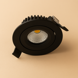 Zwarte ronde LED inbouwspot met koelribben en veerclip, schuin zijaanzicht op een beige achtergrond.