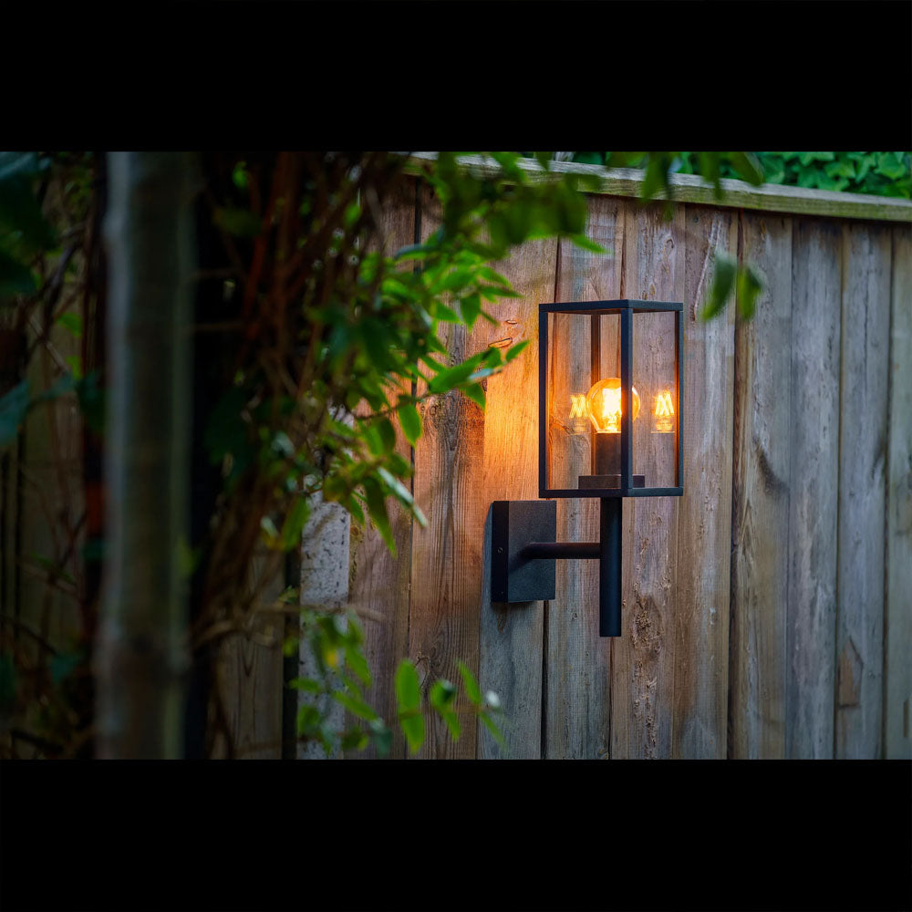 Zijaanzicht van een moderne zwarte buitenwandlamp met retro filamentlamp, gemonteerd op een houten schutting en omgeven door groenblijvende planten, verlichtend in schemerlicht voor sfeervolle tuinverlichting.