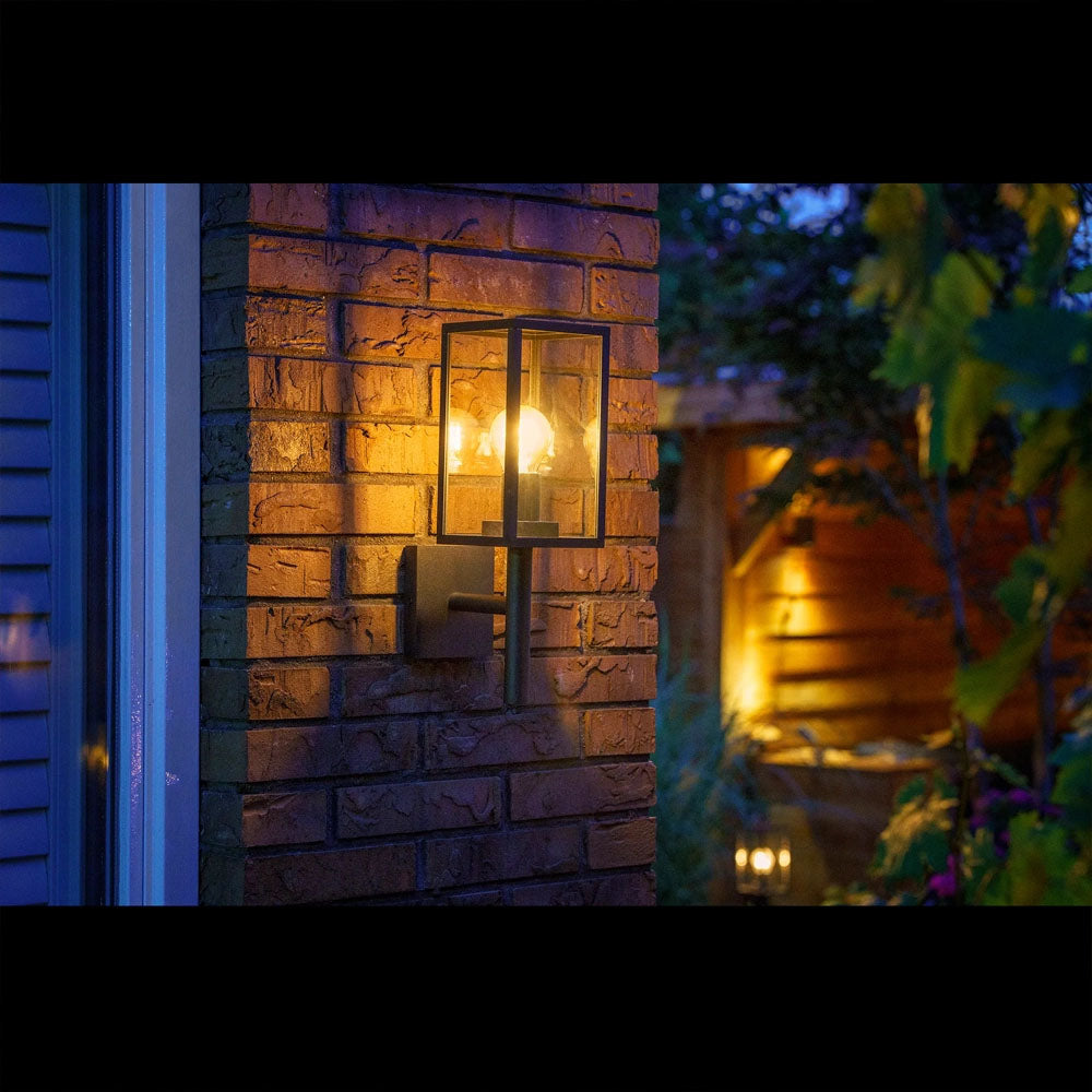 Moderne zwarte wandlamp met heldere glazen kap gemonteerd op een bakstenen buitenmuur in schemerlicht, vooraanzicht tijdens de avond in een sfeervol verlichte tuinomgeving.