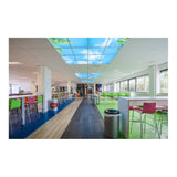 Moderne bedrijfs-kantine met kleurrijke stoelen en tafels, gezien vanuit een diagonaal vooraanzicht, met opvallend verlichte systeemplafondpanelen die een blauwe lucht met wolken en boomtoppen afbeelden.
