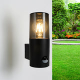 Zwarte moderne buitenwandlamp met sensor en heldere glazen cilinderkap, brandend aan een witte muur met tuin op de achtergrond – zijaanzicht.