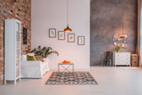 Moderne woonkamer met industriële en Scandinavische invloeden, gefotografeerd in frontaal aanzicht; witte bank met decoratieve kussens, zwart-wit geometrisch vloerkleed, houten salontafel met fruit, bakstenen muur links, betonnen accentmuur rechts, stijlvolle verlichting en wanddecoraties.