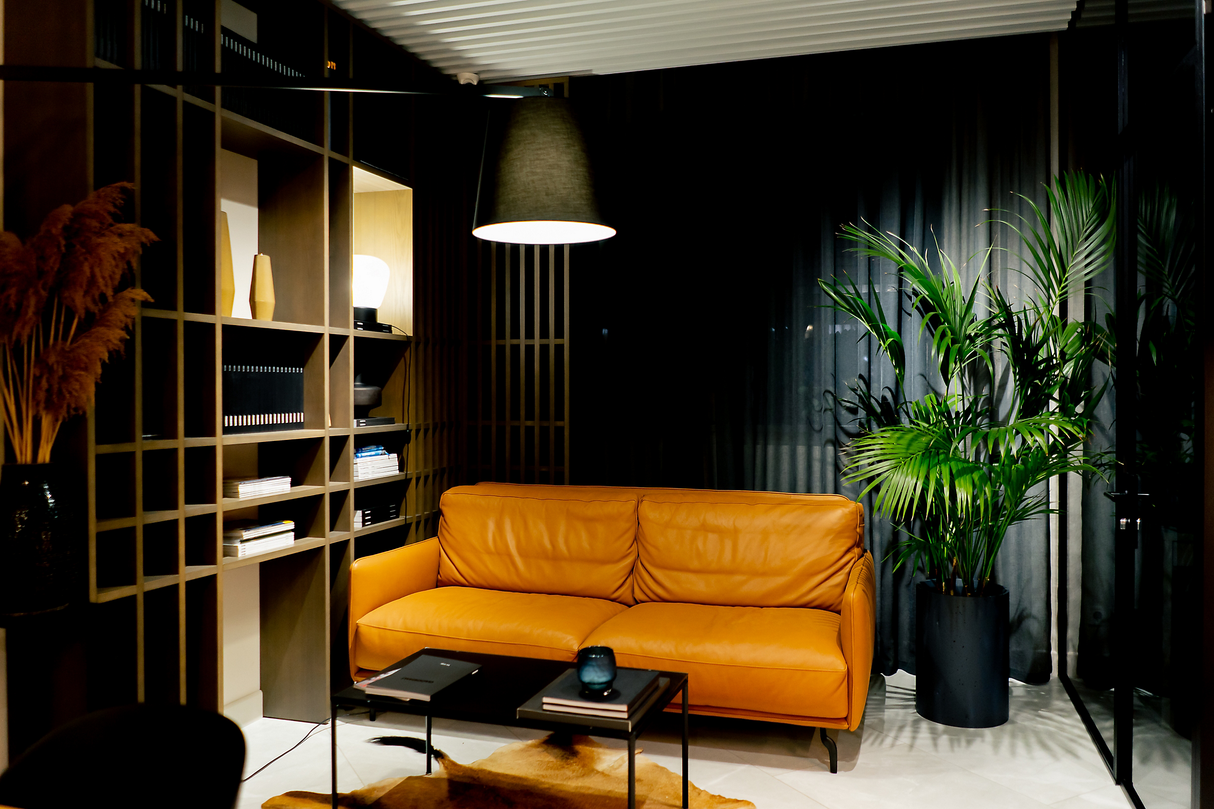 Moderne woonkamer met een frontaal aanzicht van een okergele leren designbank, geplaatst voor donkere gordijnen naast een grote groene kamerplant; links een open wandkast met decoratieve objecten en boeken, belicht door sfeervolle hanglamp.