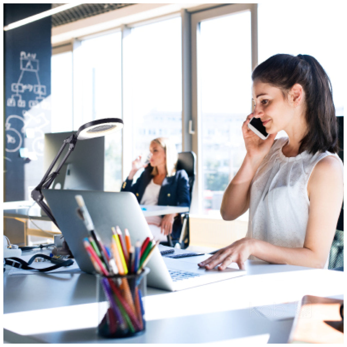Zakenvrouw aan het telefoneren terwijl ze werkt op een laptop in een modern, zonovergoten kantoor; zijaanzicht met focus op de vrouw op de voorgrond en een collega op de achtergrond.